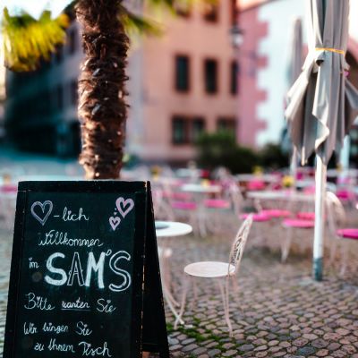 Einladungstafel „Herzlich Wilkommen im Sam's“ vor einer Palme, Café-Terrasse im Hintergrund